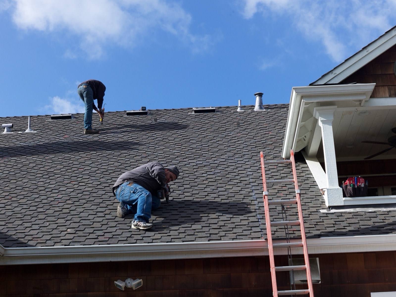 Two workers on a dark laminated shingle roof above cedar-shingled walls, one kneeling at a bracket near the eave, a ladder against the gutter