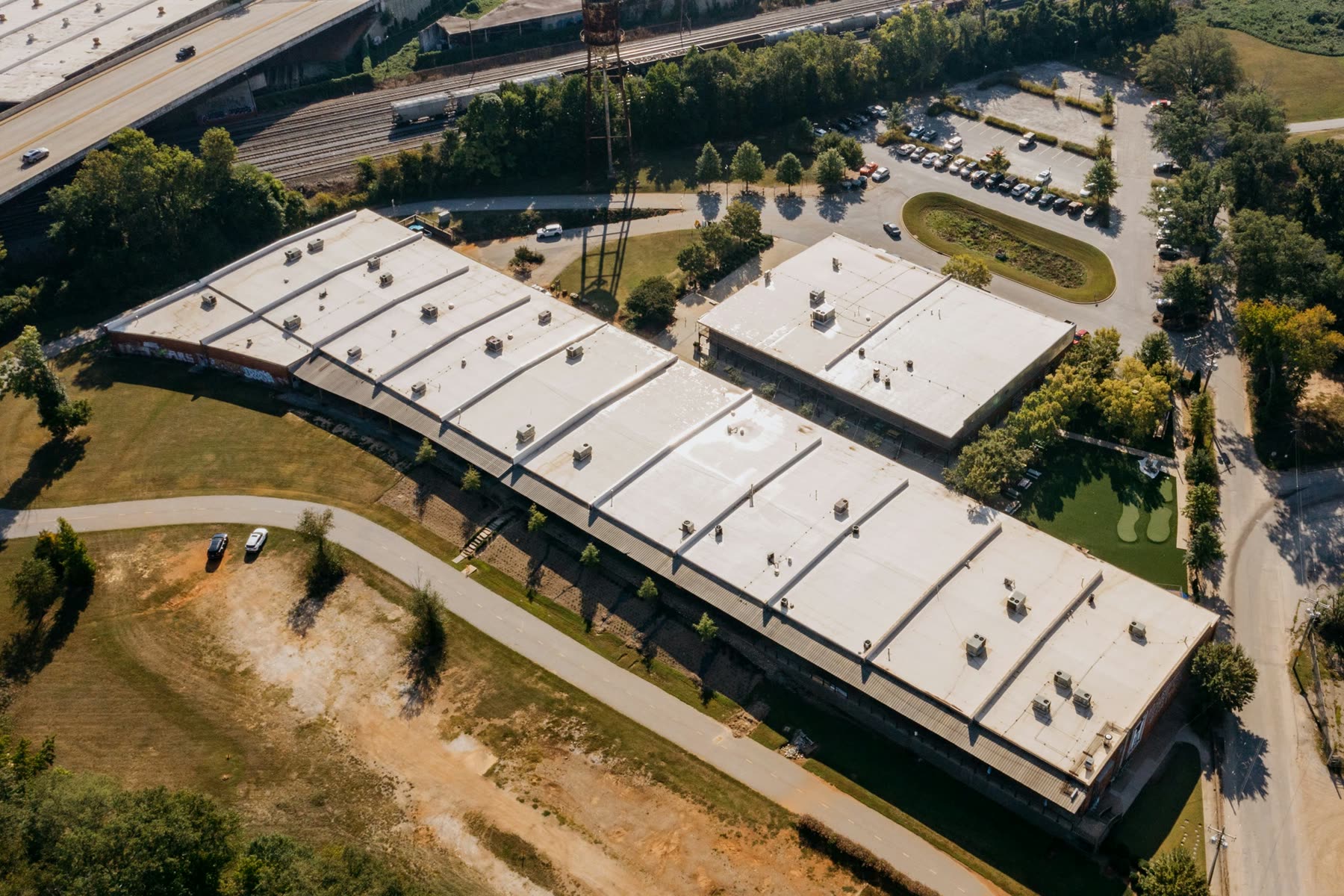 Aerial view of a long light-industrial building and a smaller shop building, both with pale low-slope roofs and rooftop units, surrounded by trees