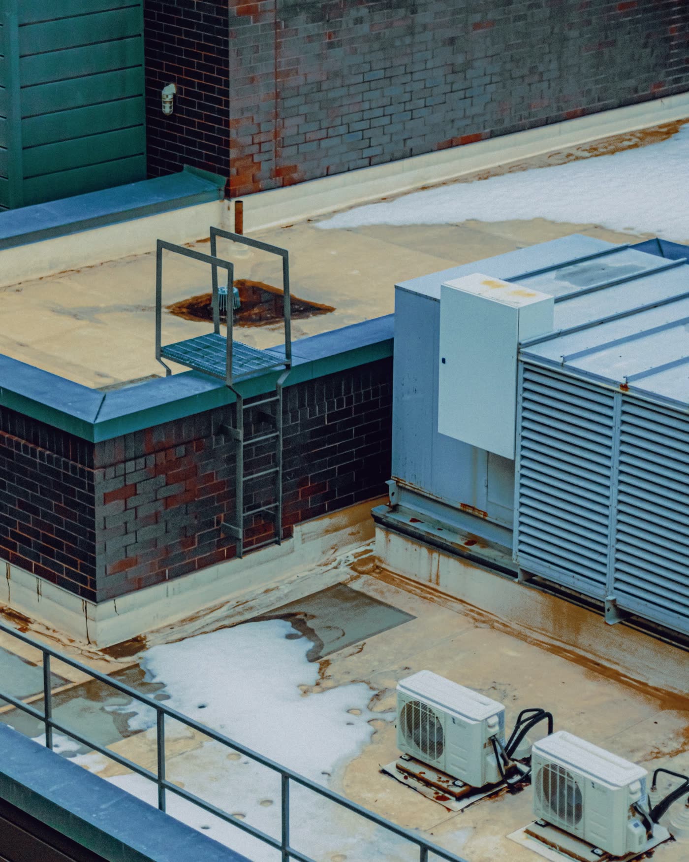 Low-slope roof with a large rooftop unit on a curb, a brick parapet with metal coping, a ladder, a roof drain with staining around it and two small condensers