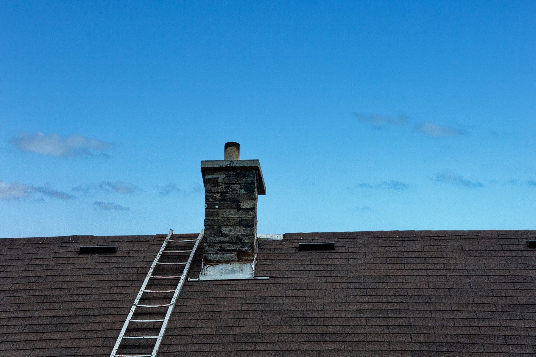 An aluminum ladder laid up a brown shingle roof beside a brick chimney, under a blue sky
