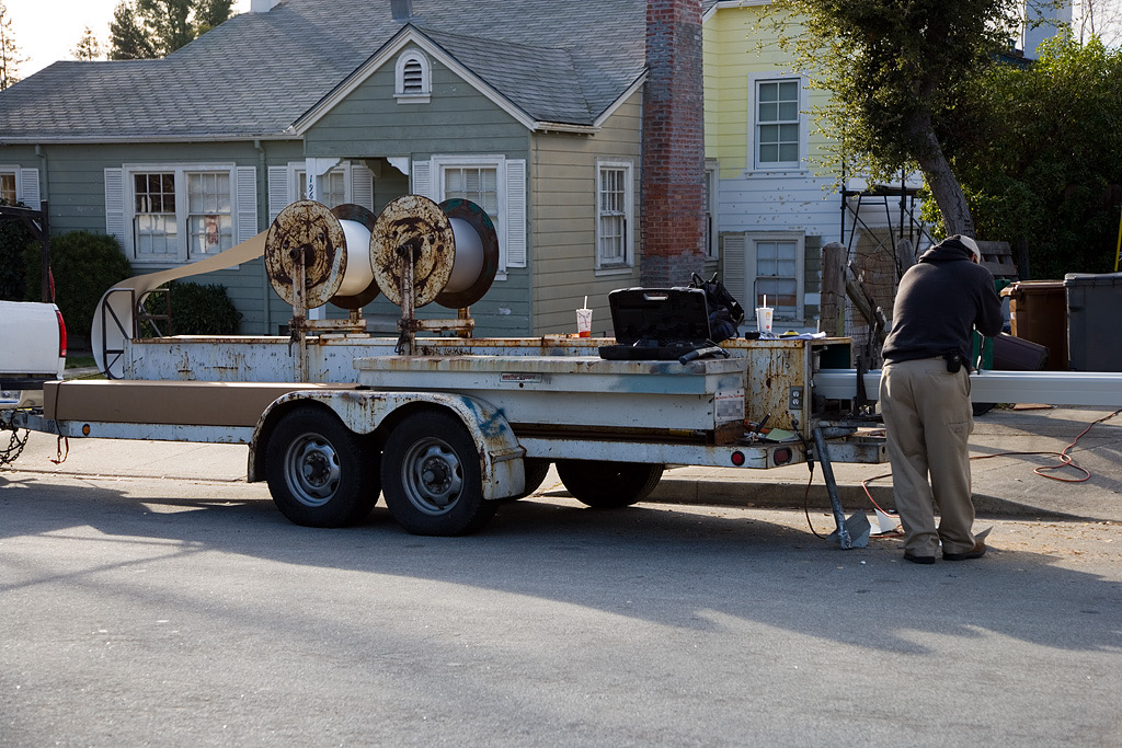 A gutter installer at a trailer-mounted seamless gutter machine parked on a residential street, with coils of aluminum on the trailer
