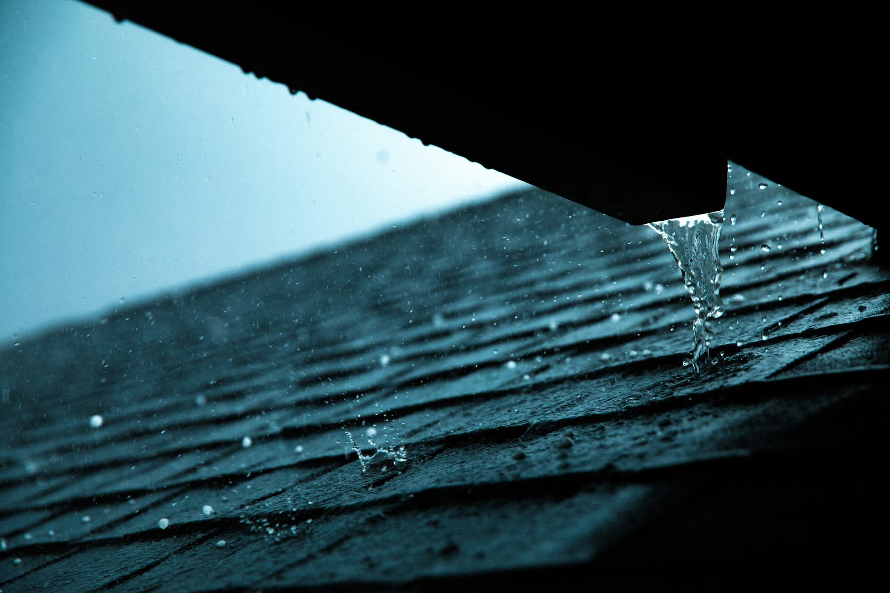 Rain pouring off the edge of an upper roof onto dark shingles below