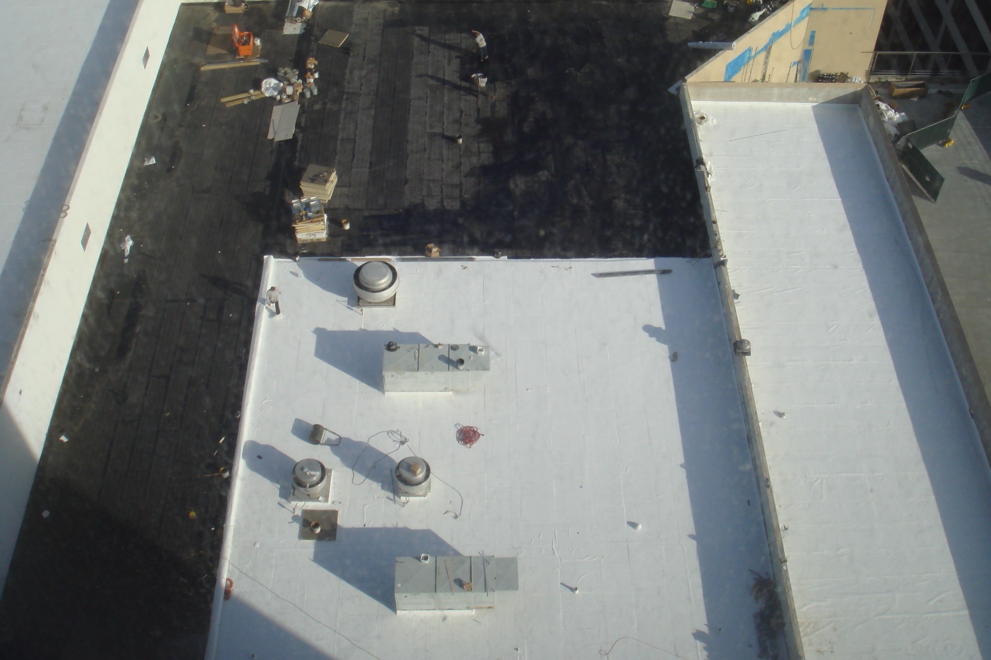 Looking down on a low-slope roof mid-job: new white membrane over the near sections, turned up the parapet walls and around exhaust fans and equipment curbs, with the old black roof still showing beyond