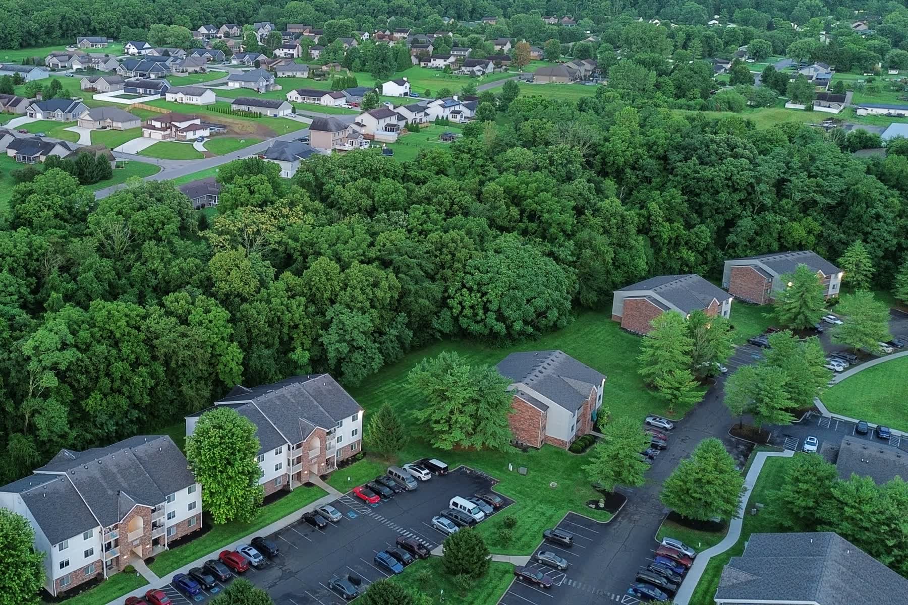 Aerial view of garden-style apartment buildings with gray shingle roofs, parking lots and tall trees
