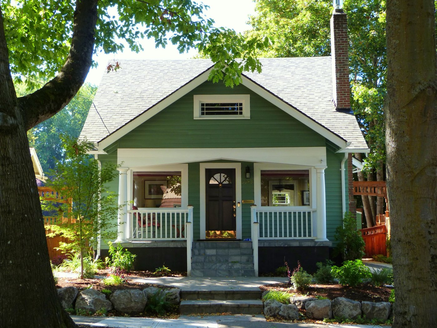 Green bungalow with a light gray shingle roof, shaded by big street trees and a neighbor's conifers